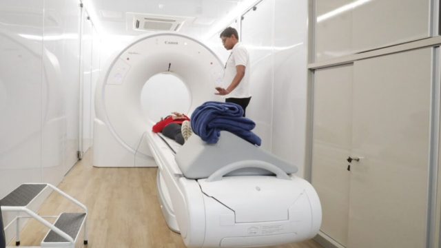 Paciente em equipamento de tomografia com profissional ao lado.