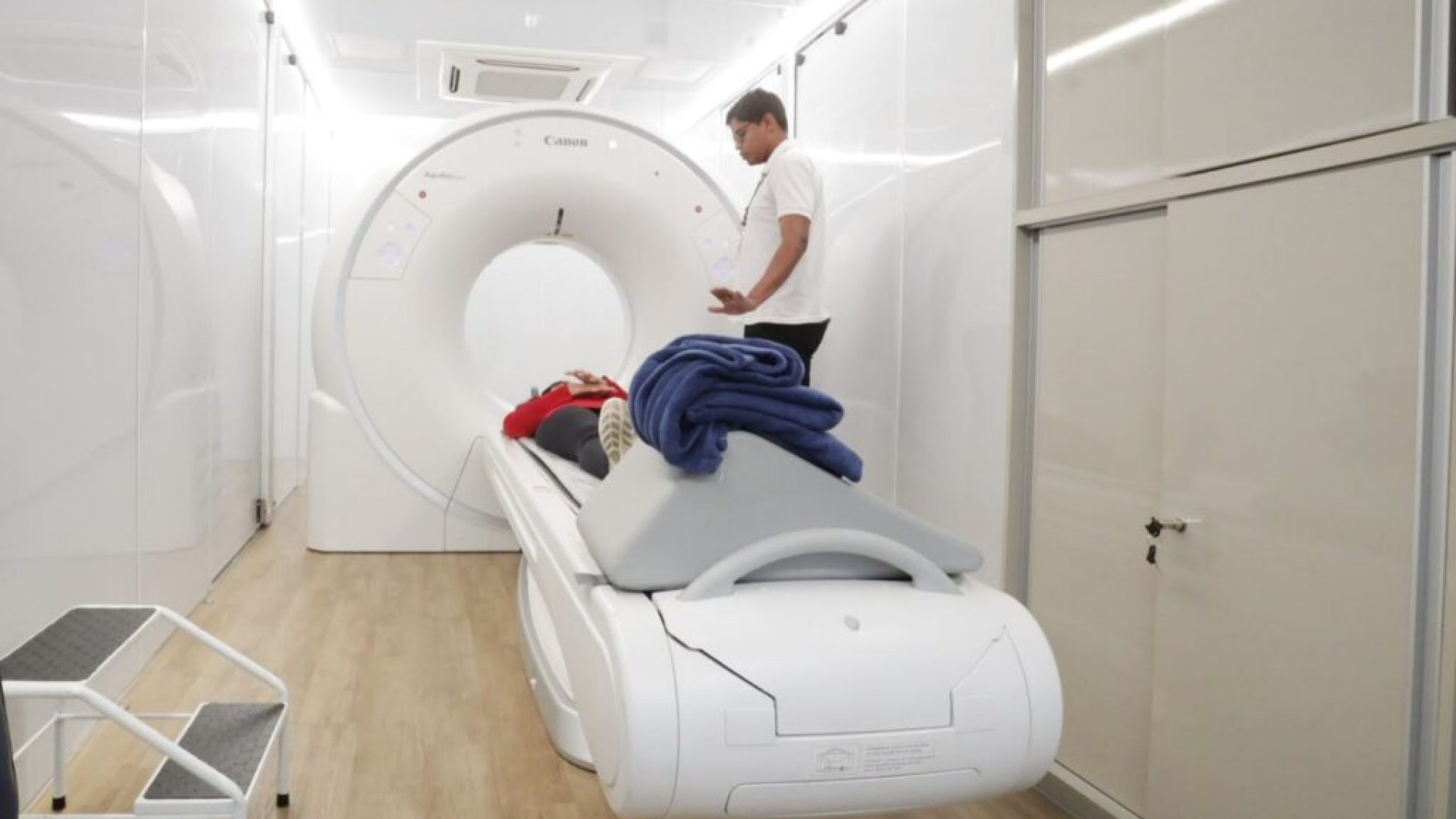 Paciente em equipamento de tomografia com profissional ao lado.
