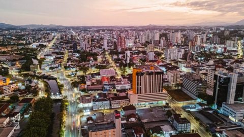 Vista aérea de Joinville ao entardecer, com ruas iluminadas e prédios.
