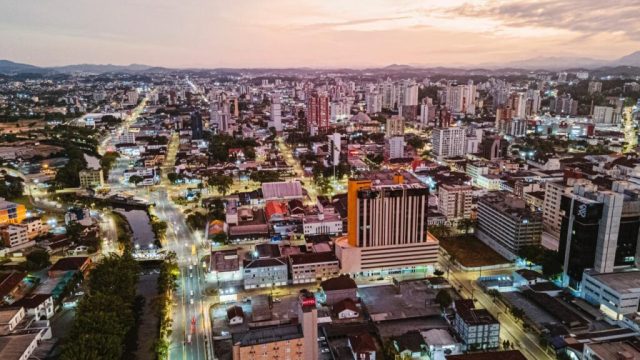 Vista aérea de Joinville ao entardecer, com ruas iluminadas e prédios.